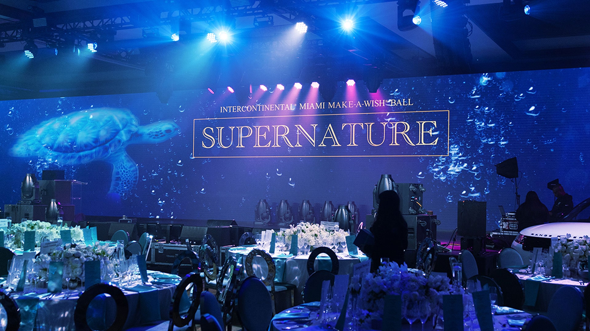 Salle de bal décorée avec des tables. L’arrière-plan de la scène montre une tortue de mer et le texte « InterContinental Miami Make-A-Wish Ball: Supernature » sous un éclairage bleu dramatique.