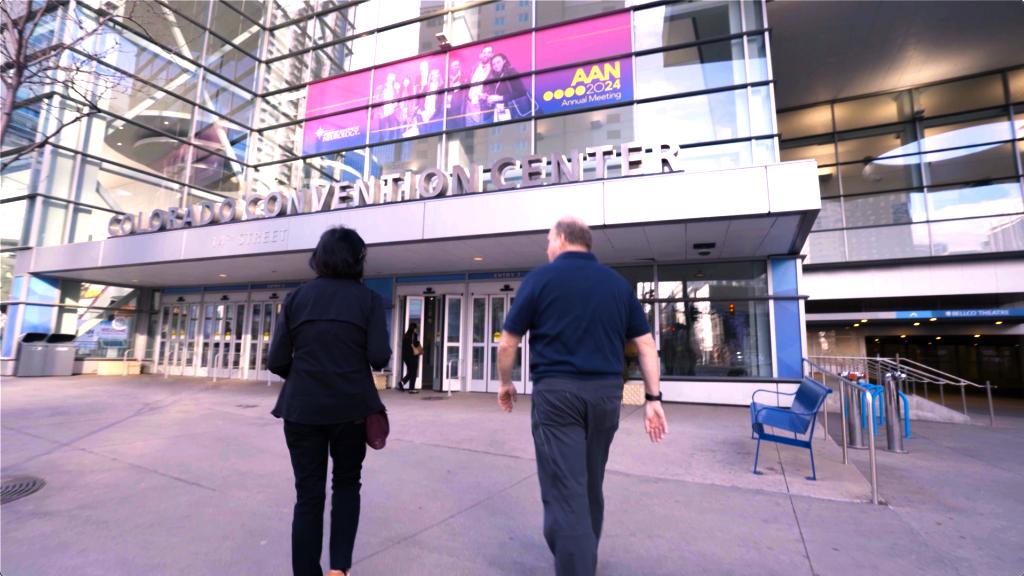 Deux personnes marchent vers l’entrée du Colorado Convention Center. Une grande bannière au-dessus annonce l’AAN 2024 Annual Meeting. Le bâtiment présente des murs de verre et des accents métalliques.
