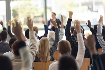 A group of people seated in a conference room raise their hands, participating in a discussion or voting, with a speaker standing in front of them.