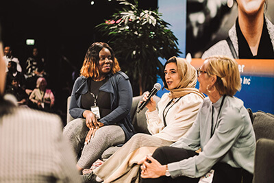 Three women sit on a panel discussion stage. One woman, wearing a headscarf, speaks into a microphone. The other two women listen attentively. There are plants and a screen in the background.