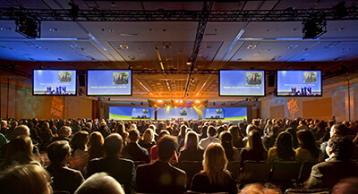A large audience sits facing a brightly lit stage with multiple large screens displaying images and text during a conference or presentation in a spacious convention hall.