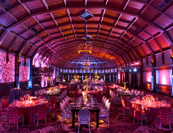 A grand ballroom with a vaulted wooden ceiling is set for an elegant event, featuring round and rectangular tables with pink and purple lighting, floral centerpieces, and elaborate chandeliers.