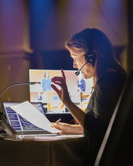 A woman wearing a headset speaks into a microphone while holding a document at a desk with a laptop and several monitors displaying people on video calls.