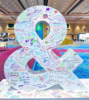 A large white ampersand sculpture covered in colorful handwritten messages and signatures stands in a spacious, brightly lit convention center with patterned carpet and tables in the background.
