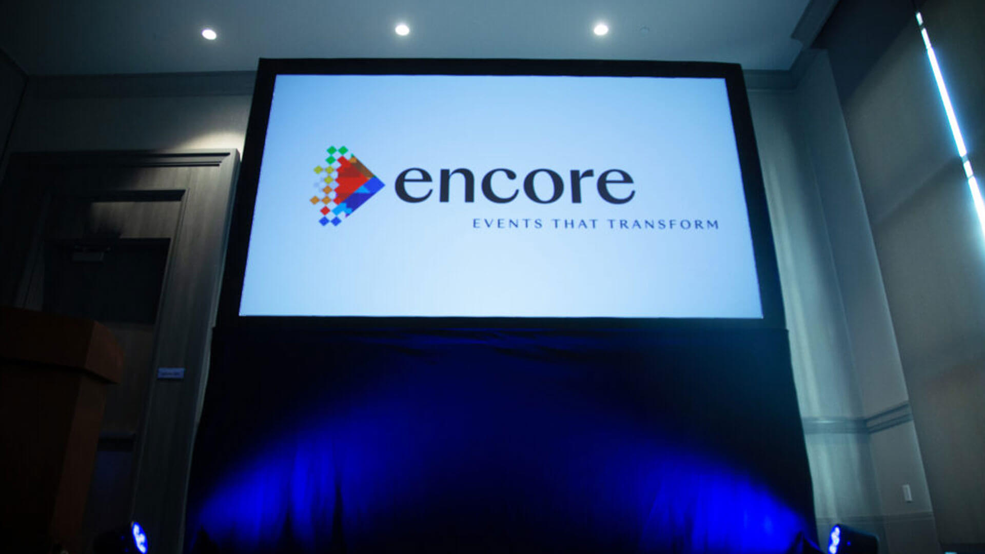 A large screen displays the Encore logo with the tagline Events That Transform in a dimly lit conference room. Blue lighting illuminates the area beneath the screen.