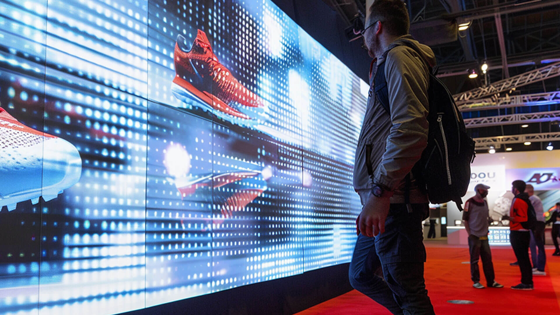 A man stands in front of a large digital screen displaying bright, colorful images of red running shoes at an indoor event or exhibition, with other attendees visible in the background.