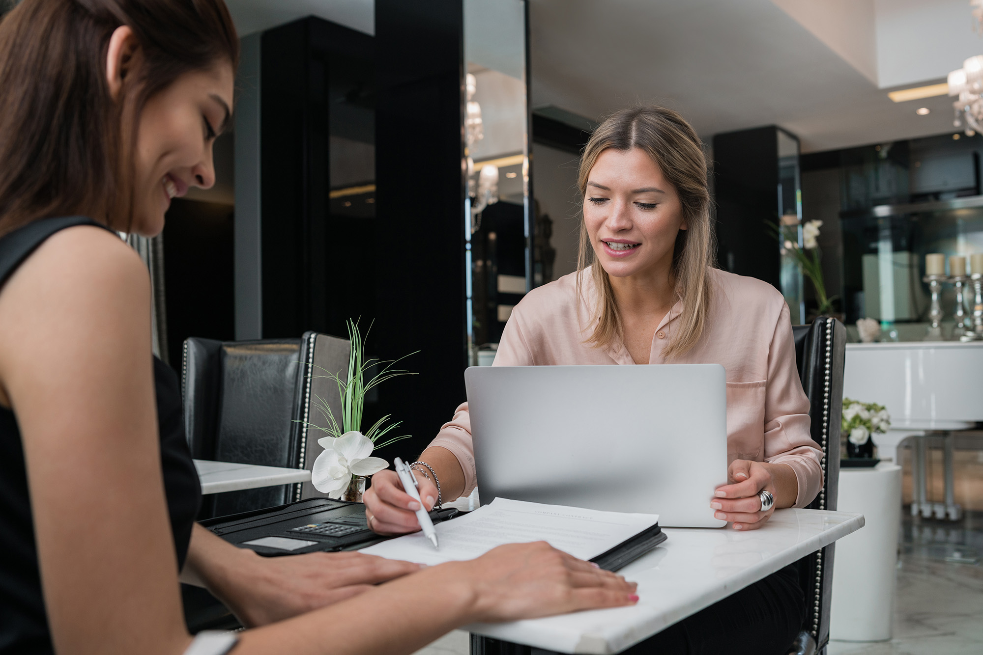 Two women sit at a table in a modern office. One woman is writing on a document while the other, with a laptop, smiles and seems to explain something. A small vase with white flowers is on the table.