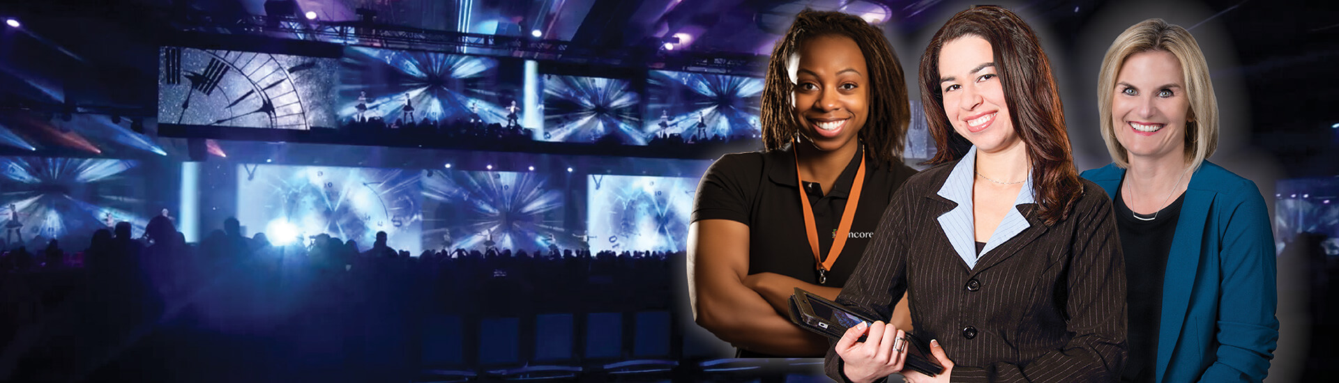 Three women stand smiling in front of a vibrant conference or event stage filled with colorful lights and a large crowd. The women appear professional, one in a polo shirt and two in business attire.