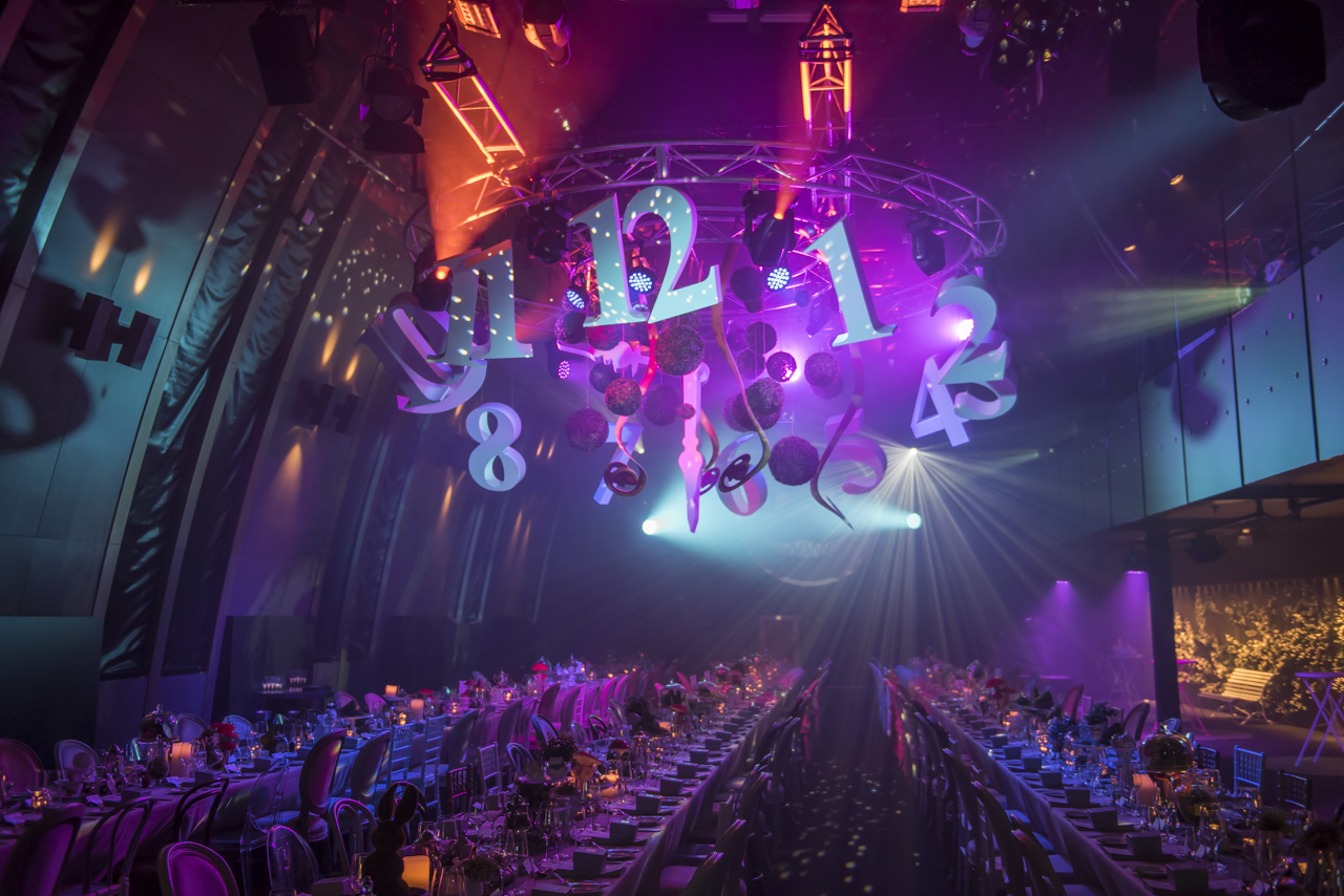 A glamorous event hall with long banquet tables, dramatic purple and blue lighting, and oversized clock numbers hanging from the ceiling, creating a festive, surreal atmosphere.