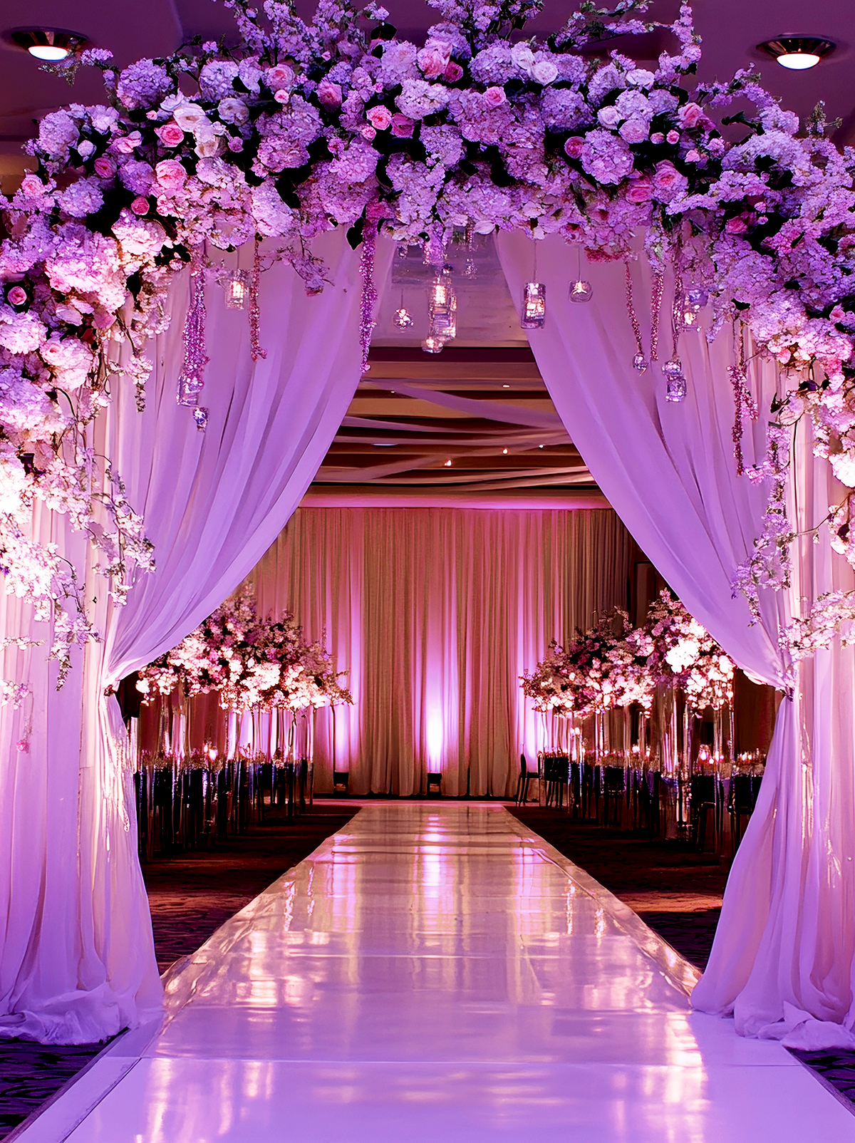 A wedding aisle decorated with a shiny white floor, tall floral arrangements, and an archway of pink and purple flowers with draped white fabric, all lit with soft purple lighting.