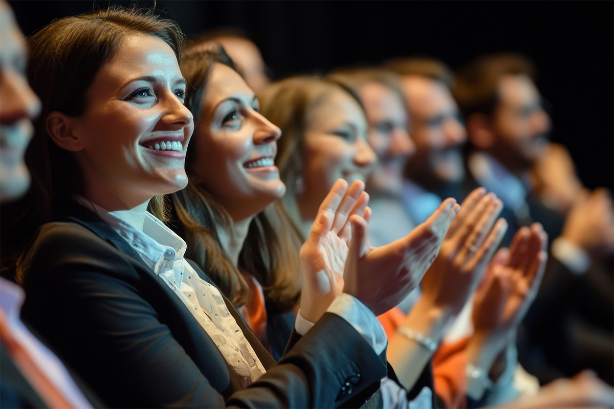 A group of professionally dressed people sit in a row, smiling and clapping enthusiastically, as if watching a presentation or performance in a dimly lit setting.