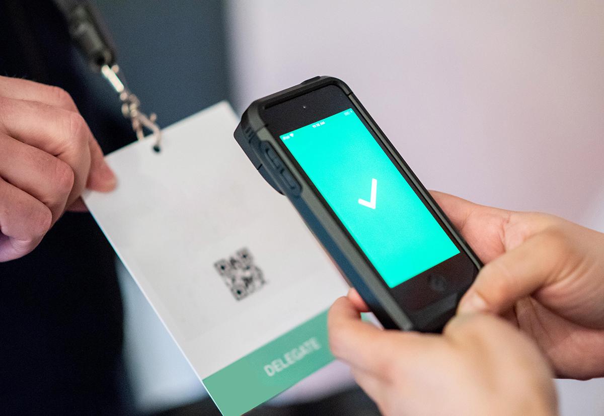 A person scans a delegate badge with a QR code using a smartphone, displaying a green screen with a white check mark, indicating successful verification.