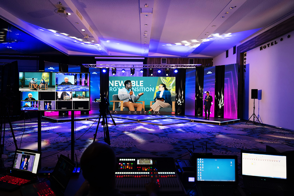 A brightly lit stage features two speakers seated and talking for a live event. Multiple screens and cameras show various angles, with control equipment and computers set up in the foreground.
