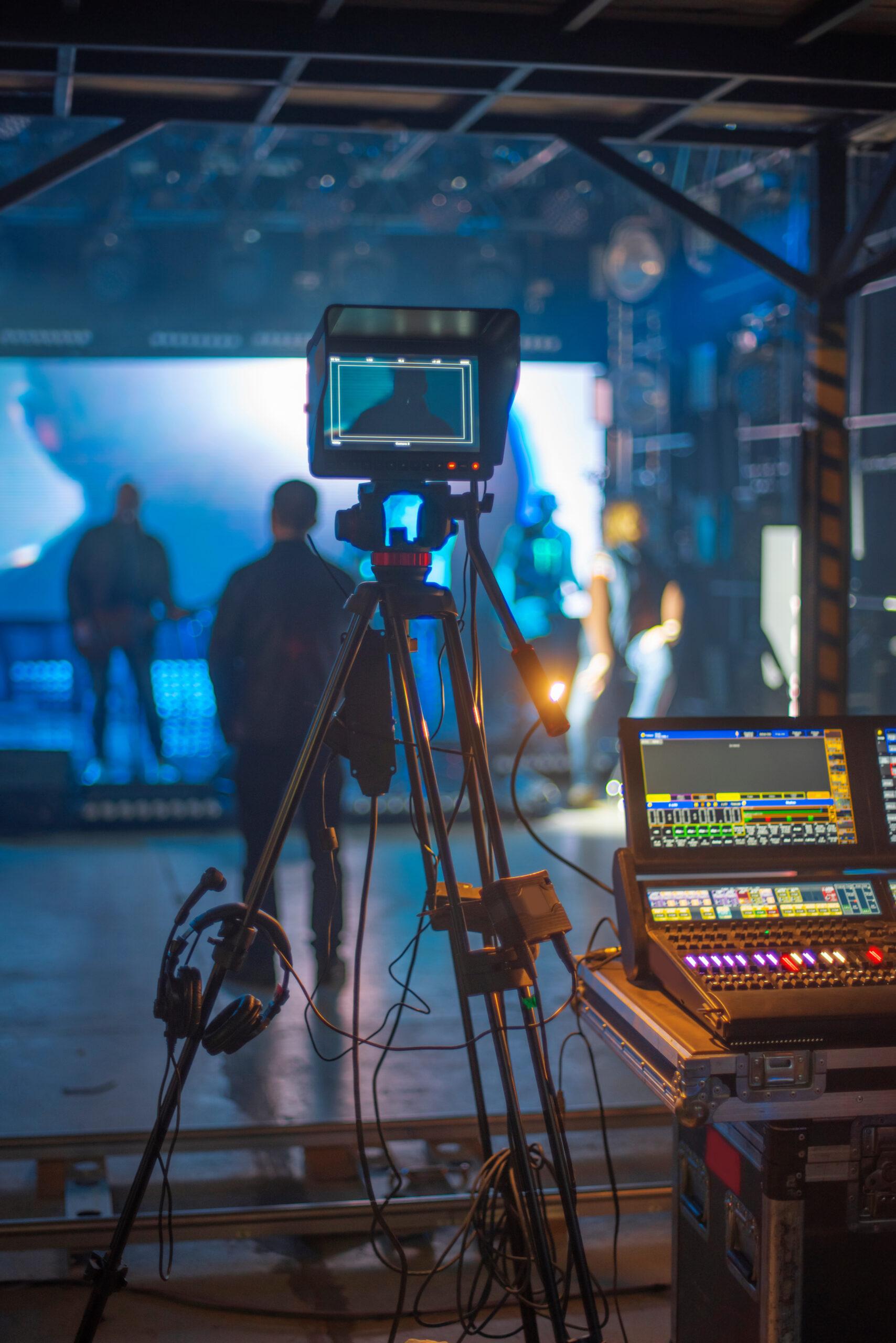 A video camera on a tripod is set up in a studio with stage lights, sound equipment, and a control panel; several people are visible in the blurred background, preparing for a performance or recording.