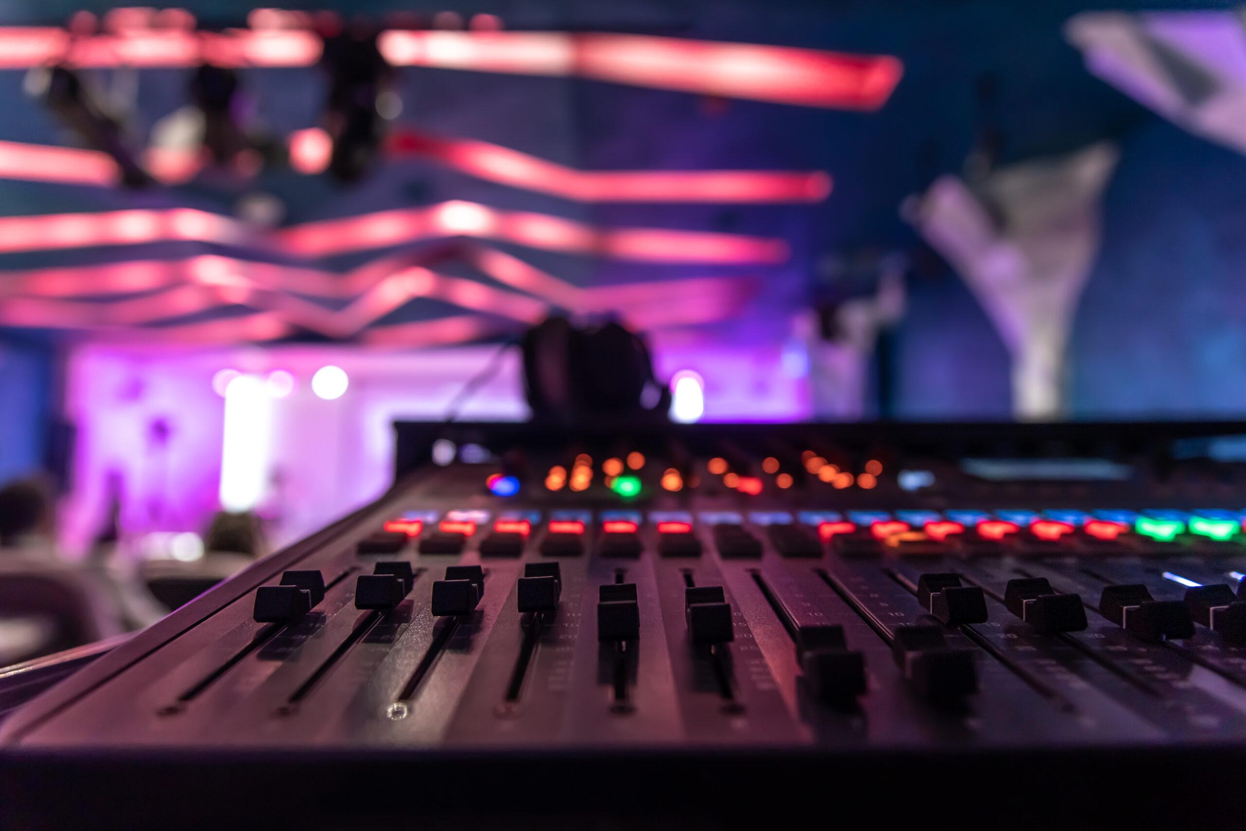 A close-up of an audio mixing console with colorful lights, sliders, and buttons in focus. The background shows a blurred stage with pink and purple lighting and modern ceiling décor.