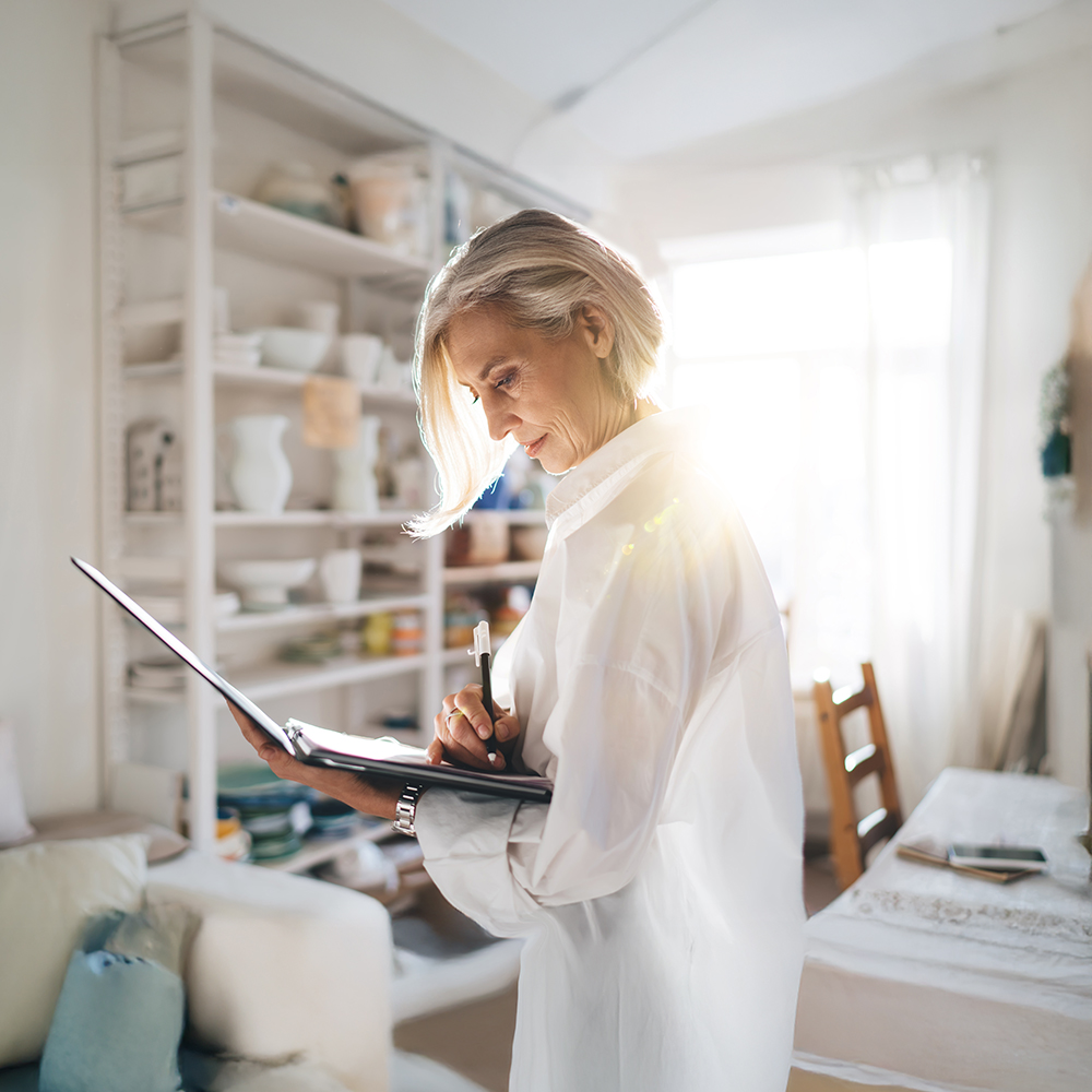 An older woman with gray hair stands in a sunlit room, writing on a clipboard. She appears focused, dressed in a white shirt, with shelves and furniture visible in the bright, cozy background.
