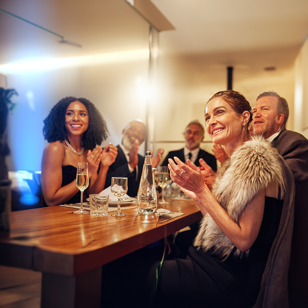 A group of elegantly dressed people sit around a wooden table with drinks, smiling and applauding in a warmly lit, upscale restaurant or event setting.