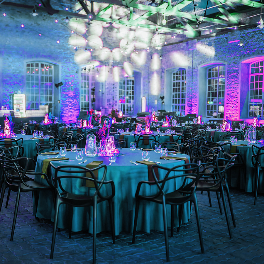 A banquet hall with round tables covered in teal cloths, set with glassware and lit by vibrant purple and blue lighting. Modern black chairs surround the tables, and the room features large windows and exposed brick walls.