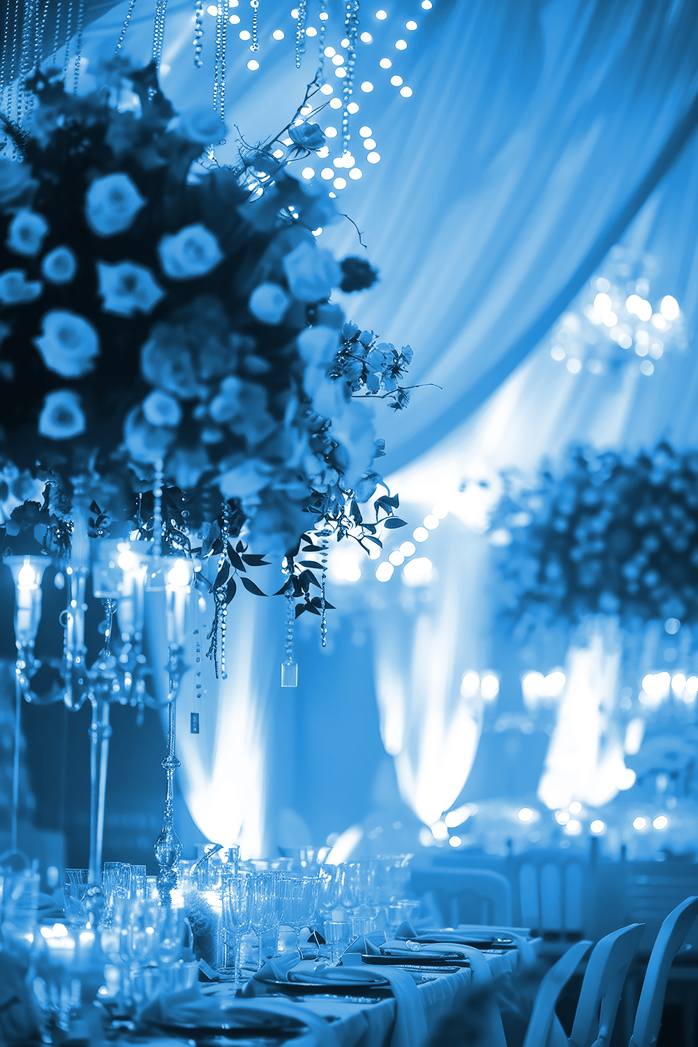 Elegant event hall decorated with draped fabric, hanging lights, and large floral centerpieces on tables set with glassware and candles, creating a luxurious and festive atmosphere with a blue color tone.