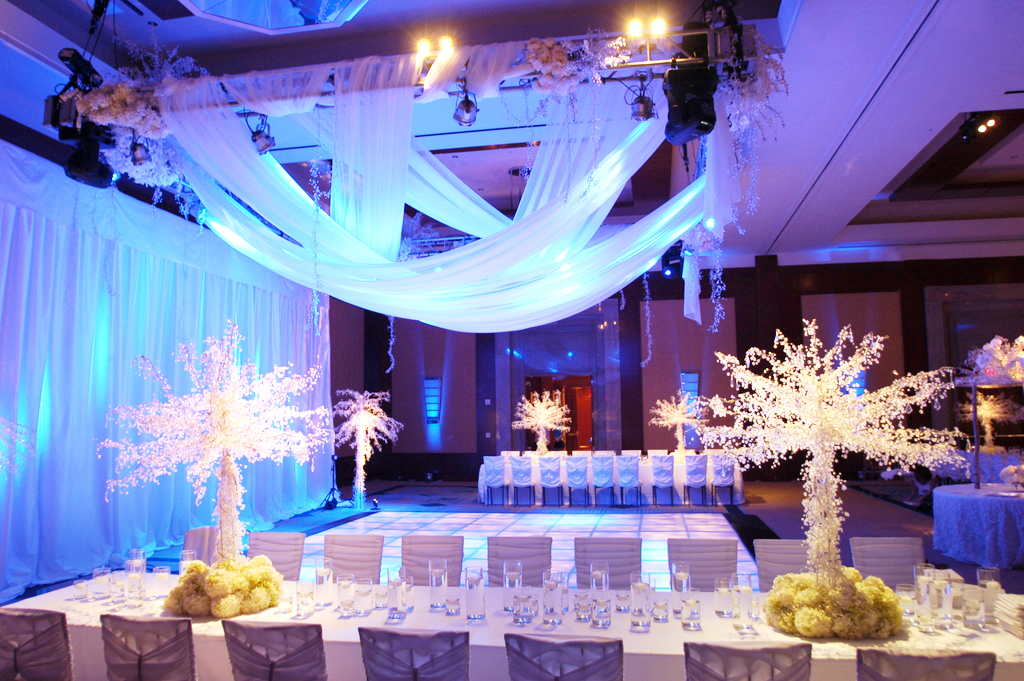 A lavishly decorated banquet hall features white draped fabric, glowing blue uplighting, ornate white floral centerpieces, and elegant table settings around a central dance floor.