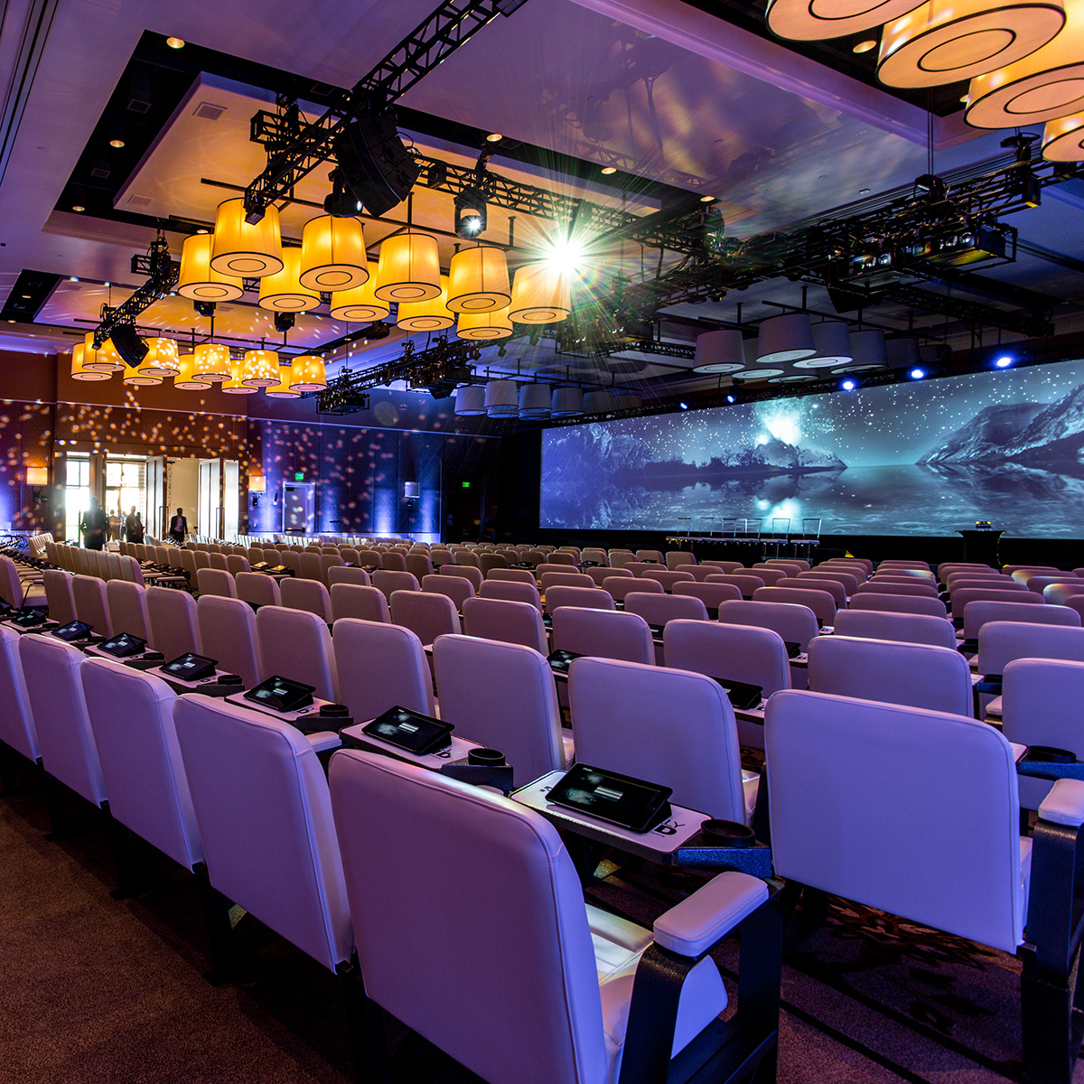 Rows of white chairs face a large stage with multiple screens displaying a blue cosmic scene in a modern, spacious conference hall with dramatic lighting and elegant hanging lights.