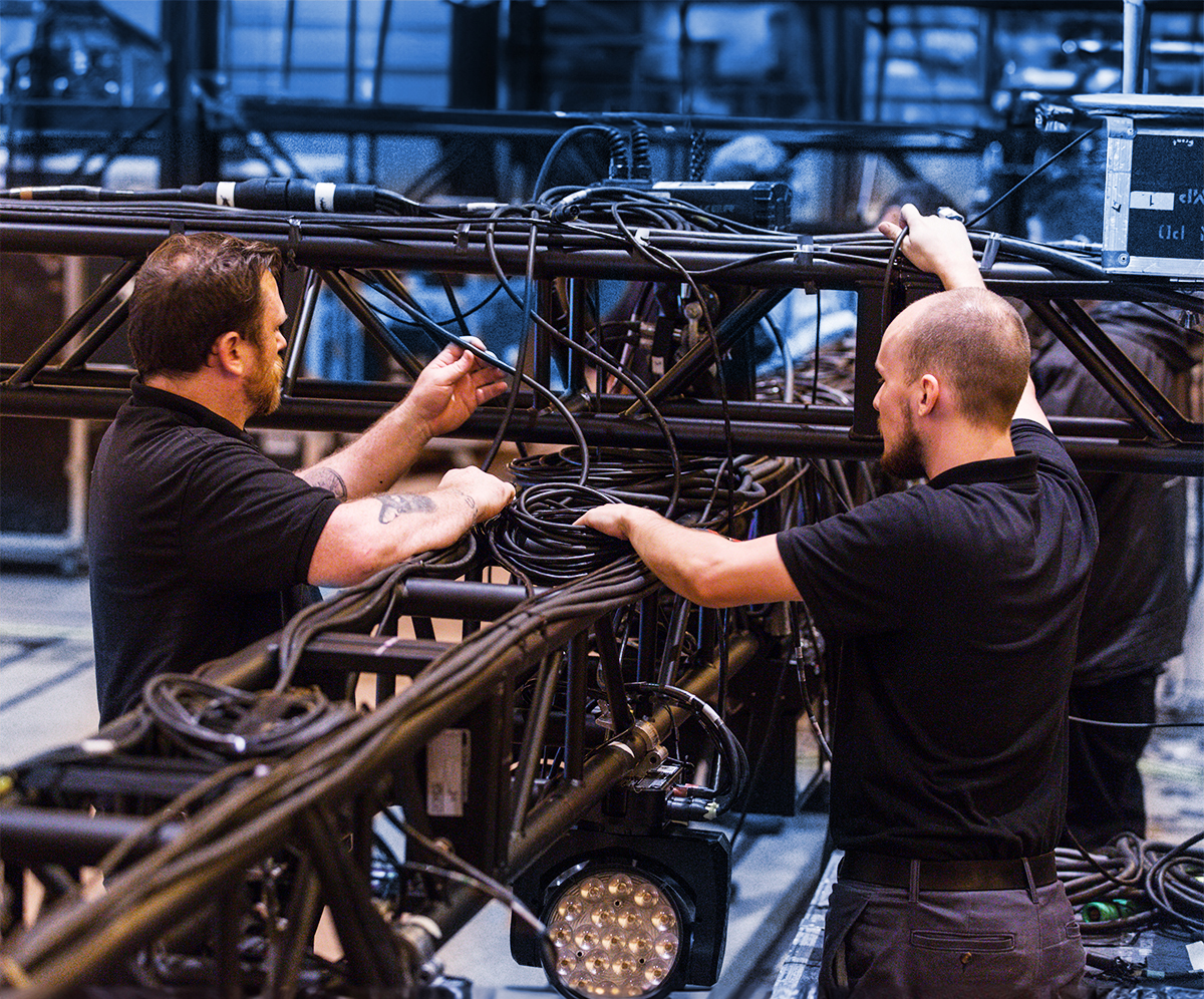 Two men in black shirts work on setting up stage lighting equipment, adjusting and organizing cables on a metal truss structure in an indoor venue.