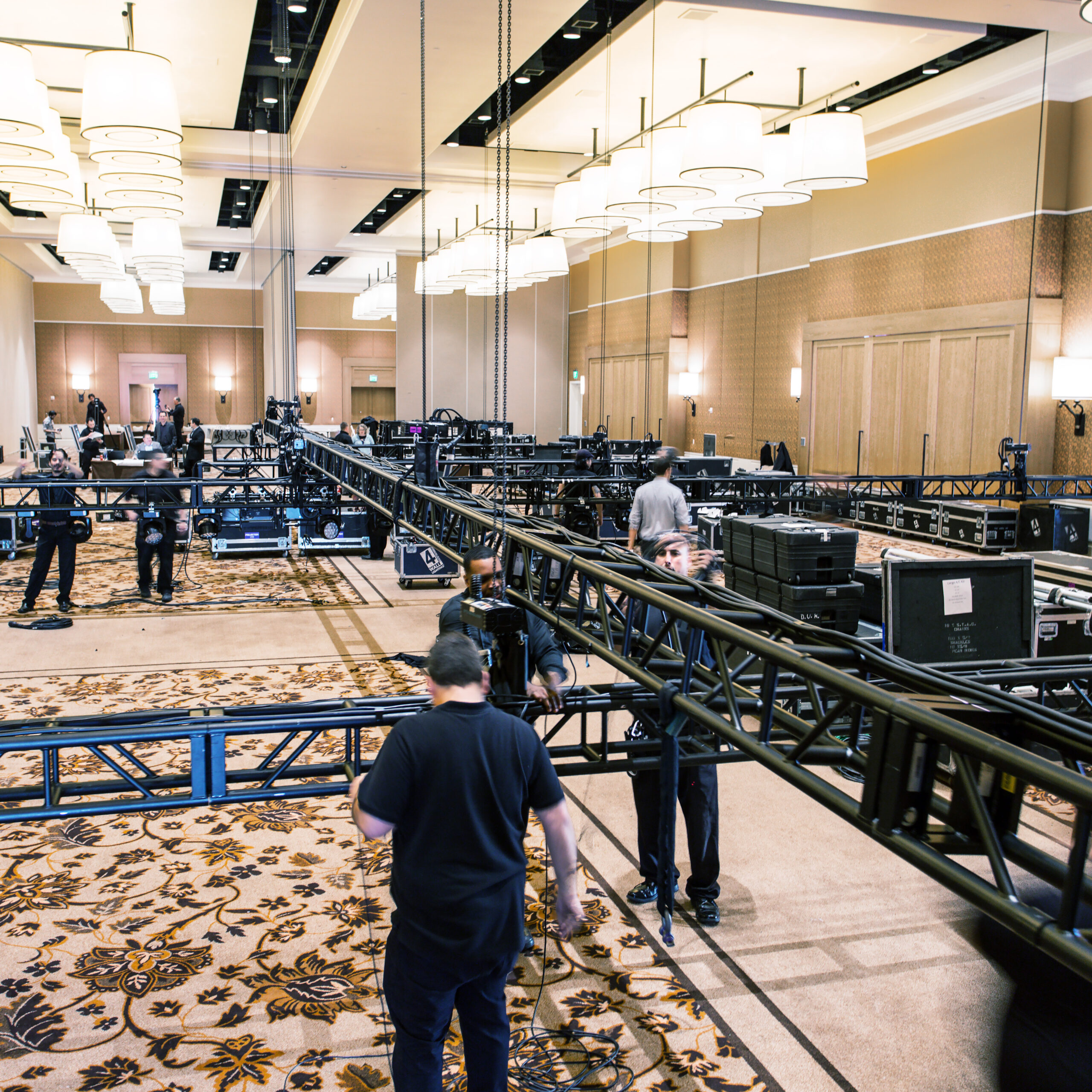 Several workers set up metal trusses and equipment in a large, carpeted ballroom with high ceilings and hanging lights, preparing the space for an event or production.