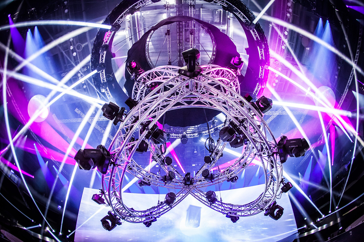 A large, circular metal lighting rig with spotlights hangs from the ceiling, surrounded by colorful purple and white beams of light in a dark concert or event venue.