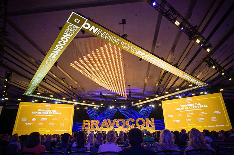 A large, brightly lit BravoCon stage with yellow signage reads “BRAVOCON” and “Bringing the Miami Heat to Vegas.” Audience members sit facing a panel of speakers under a decorative ceiling display.
