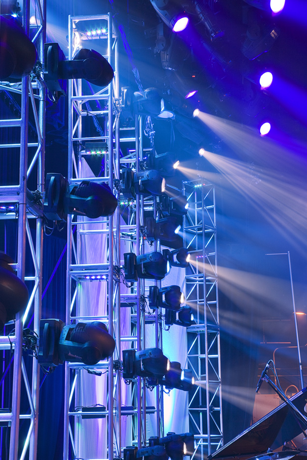 Rows of stage lights mounted on metal trusses shine blue and white beams across a stage, creating a vibrant and dramatic atmosphere for a live performance or event.