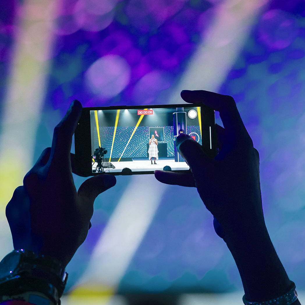 A person holds up a smartphone, capturing a photo or video of a speaker on stage under bright lights with a colorful, blurred background.