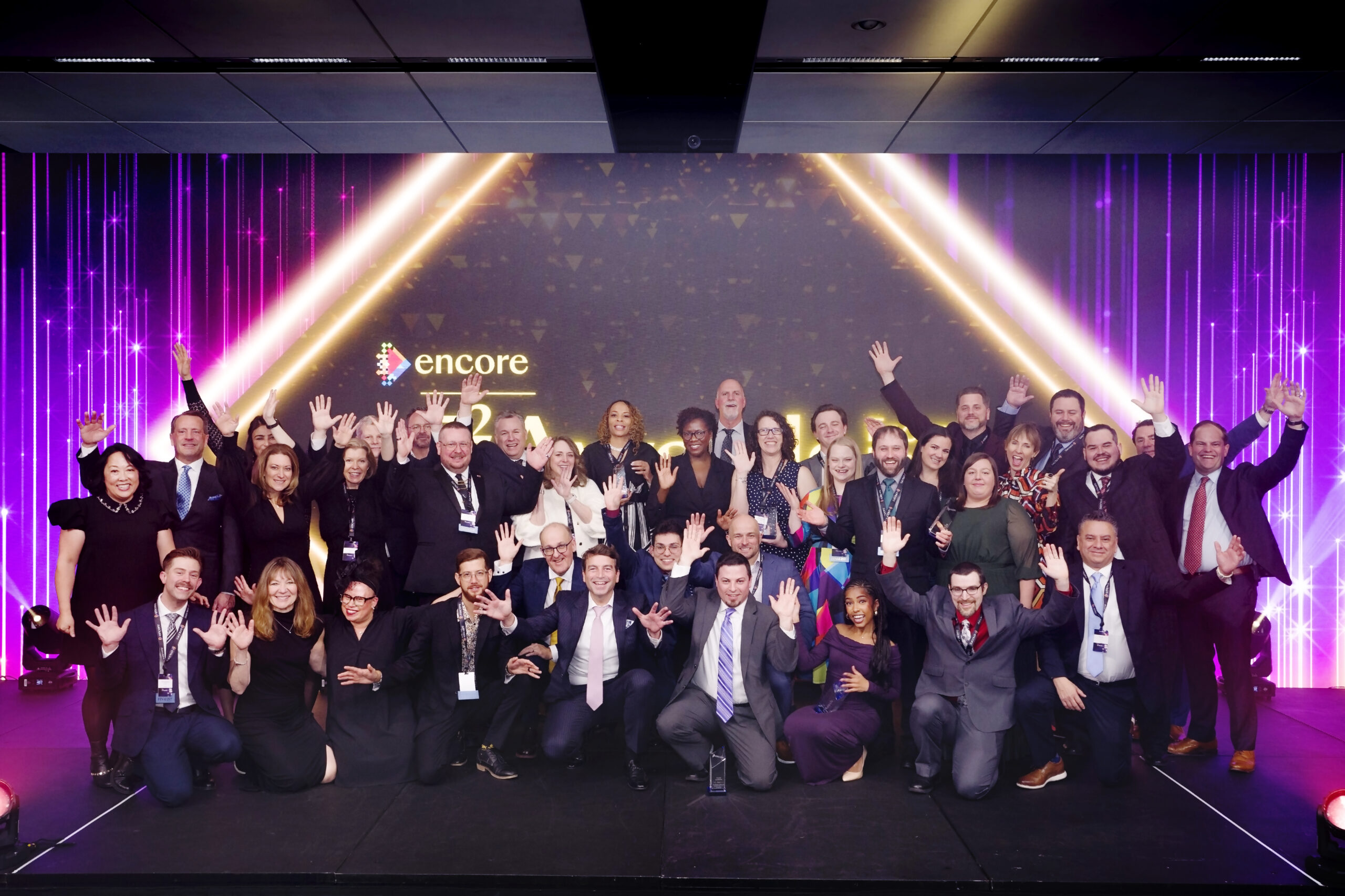 A large group of people in formal attire pose together on a stage, smiling and waving with hands raised. Behind them is a colorful, illuminated backdrop with the word encore and a geometric light display.