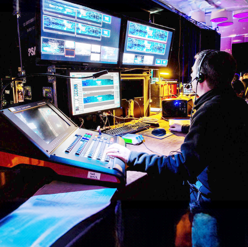 A person wearing a headset operates a control panel and several monitors in a dimly lit control room, managing video feeds and live event production.