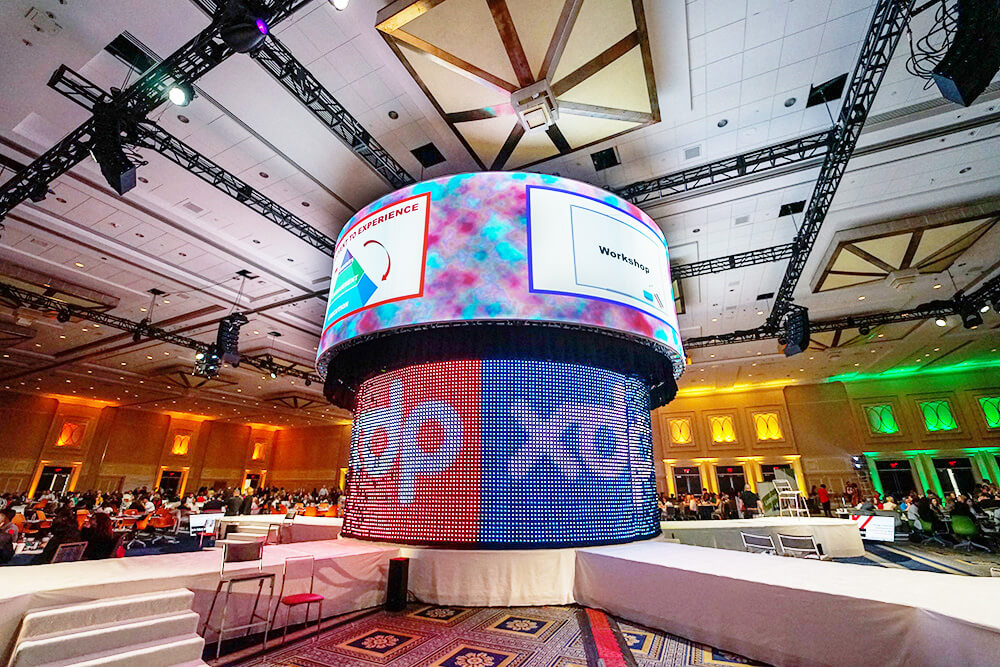 A large, circular digital display with colorful lights and graphics is suspended in the center of a spacious conference hall filled with attendees seated at round tables. The room is lit with multicolored uplighting.