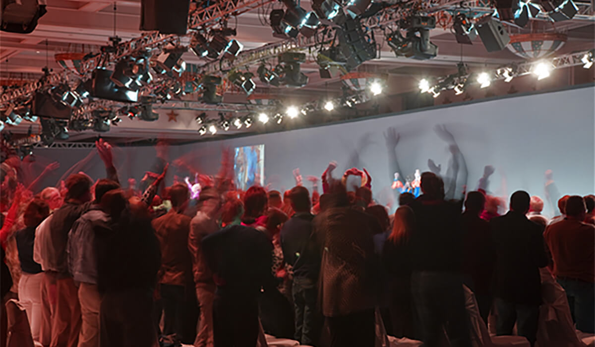 A large audience stands and cheers in front of a brightly lit stage at an indoor event, with raised hands and blurred motion indicating excitement. Stage lights and equipment hang overhead.