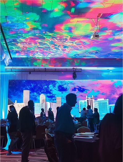 People gather in a large event room with colorful, swirling lights projected on the ceiling and a city skyline backdrop on the wall, creating a vibrant, lively atmosphere. Silhouettes of attendees are visible in the foreground.