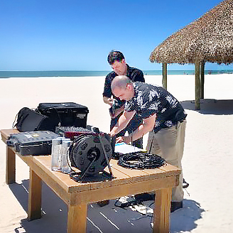 Two men set up audio equipment on a wooden table on a sunny beach with white sand. A thatched-roof hut and the ocean are visible in the background under a clear blue sky.