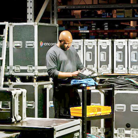A man stands in a warehouse surrounded by large black equipment cases, organizing a bundle of cables on a cart with yellow trays. Shelves with various items are visible in the background.