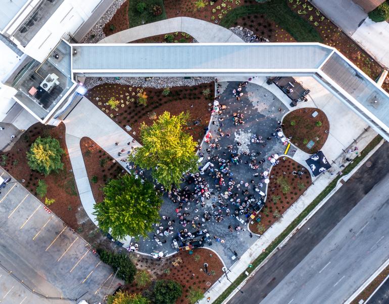 Aerial view of a crowd gathered in a landscaped outdoor area near a large building, with pathways, greenery, and a covered walkway crossing above. Cars are parked along a nearby street and parking lot.